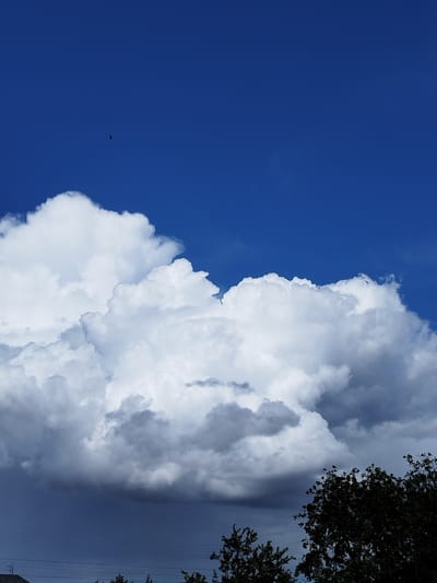This is a vertical, eye-level, outdoor, daytime photograph of a bright blue sky with large, white, puffy cumulus clouds dominating the middle of the frame. In the upper portion of the sky, a small, dark, oblong object is visible, possibly a balloon or a kite. The lower part of the image shows the dark underside of the clouds, transitioning to a darker blue sky, with the tops of trees silhouetted against it, and a faint line of utility wires. The lower left corner shows the roofline of a building. The scene depicts a cloudy day in Lozova, Ukraine.