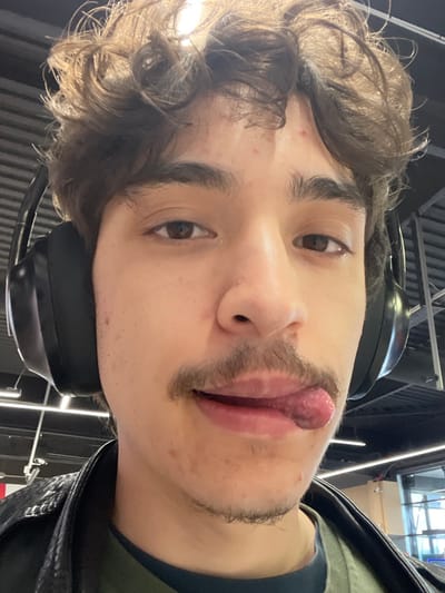 The image displays a close-up, frontal portrait of a young male individual, approximately 18-30 years old, situated indoors in Puyvert, France. He possesses curly brown hair, dark brown eyes, and a mustache. Several small red blemishes are visible on his forehead and cheeks. The individual is wearing black over-ear headphones, a dark-colored shirt, and a black leather-style jacket. His tongue is extended from the right side of his mouth, curving slightly downwards. The background is blurred, revealing an indoor setting with a high ceiling, horizontal dark paneling, and bright overhead lighting fixtures. A faint reflection on the right side suggests a window or glass partition.