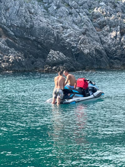 Two shirtless men are on a jet ski in the clear, turquoise waters near a rocky cliff face. One man, wearing patterned swim trunks, stands in the water next to the jet ski, facing away from the camera. The other man, wearing blue swim trunks, is seated on the jet ski, also facing away. A bright red life vest is attached to the back of the jet ski, along with a black bag. The jet ski has "YAMAHA" branding visible on its side. The rocky cliffs, characteristic of the coastline in Dubraljevina, Montenegro, form a backdrop to the scene. The water is calm with light ripples and reflections of the sun.