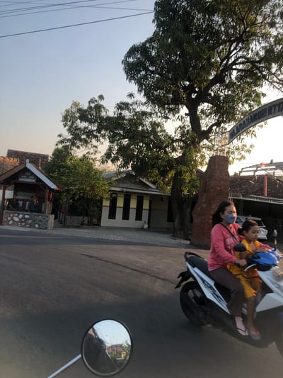 The image depicts a street scene in Glagah Arum, Indonesia, during the late afternoon or early evening. The sky is clear and a pale blue, with power lines crisscrossing the upper portion of the frame. A large tree with dense foliage dominates the middle ground, casting shadows on the surrounding area. To the left of the tree, a small, open-sided structure with a wooden roof and stone base is visible, where two individuals are seated. Adjacent to this structure is a building with a yellow and gray facade and dark windows.
In the foreground, a white scooter is being ridden by two individuals. The person in front, presumably an adult woman wearing a pink long-sleeved shirt and brown pants, has a light blue mask covering her lower face. She is driving the scooter. The person behind her, a child wearing a yellow shirt, is seated in front of her. They are both wearing flip-flops. The scooter is moving on an asphalt road.
A brick pillar and an arched sign are visible to the right of the tree, with the text "GLAGAHARUM RTT" partially visible on the sign. Further in the background, a white car is parked and a tiled roof of a building is visible. A motorcycle's circular rearview mirror is in the bottom left corner of the image, reflecting a street lamp and buildings. The overall impression is a snapshot of everyday life in a residential or semi-commercial area of Glagah Arum.