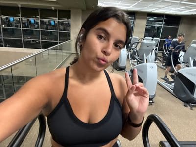 The image depicts an indoor gym environment, located in Plainsboro Township, United States, at night. In the foreground, a young adult female with dark hair, wearing a black sports bra and a white earbud in her right ear, is taking a selfie. She is looking directly at the camera with pursed lips and making a peace sign (V-gesture) with her left hand. The arm of a gym machine is partially visible below her. In the midground, several elliptical trainers are arranged. Two male individuals are present in the background; one is actively using an elliptical machine, dressed in a dark short-sleeved shirt and dark shorts. To the left, a large window overlooks a blurred nighttime landscape with distant lights, featuring a glass and metal railing. The gym floor is covered with light-colored commercial carpeting, and the space is illuminated by artificial overhead lighting.