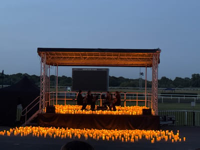 The image depicts a stage set up at Kempton Park in the Borough of Spelthorne, United Kingdom. The stage is surrounded by numerous lit candles. Four musicians are seated on the stage and appear to be performing. A large digital screen is visible behind the stage, displaying the name "KEMPTON PARK". The background includes trees and a race track. The scene is lit by the candles and stage lighting, suggesting it is dusk or evening.