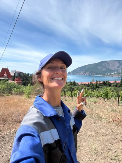 A woman wearing a blue baseball cap and a blue and grey work jacket smiles at the camera while holding up two fingers in a peace sign. Her fingers appear to be wrapped in white tape. She is standing in a dry, grassy area with grapevines in the background. In the distance, there is a body of water, a mountainside, and buildings. The sky is blue with some scattered clouds. The scene is likely in the city of Utios, Ukraine.