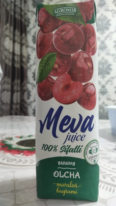The image displays a carton of "Meva juice" positioned on a table, with a patterned curtain in the background. The juice carton is predominantly white with green sections at the top and bottom. Its front face features a prominent illustration of numerous ripe, red cherries, some shown whole and some halved, all covered with visible water droplets, conveying freshness. A green leaf is also depicted among the cherries.
At the top of the carton, the brand "AGROMIR group of companies" is visible. The product name "Meva juice" is printed in a large, stylized blue font. Below this, the text "100% Sifatli" (meaning '100% quality' in Uzbek) is present. The flavor, cherry, is indicated by "Вишня" (Russian) and "OLCHA" (Uzbek) on a green band. Further down, the phrase "mevalar bayrami" (Uzbek for 'fruit festival' or 'feast of fruits') is written in yellow script. A "Halal certification body CERTIFIED" logo is also prominently featured on the carton's right side.
The table beneath the carton is covered by a light-colored tablecloth with a subtle pattern. A round coaster or placemat with a green and red pattern is partially visible on the left side of the table. The background is blurred, showing a dark, patterned curtain, suggesting an indoor setting. The image is well-lit, implying it was taken during the daytime. No individuals or specific activities are depicted, focusing solely on the juice product in a home environment in Uchquduq, Uzbekistan.