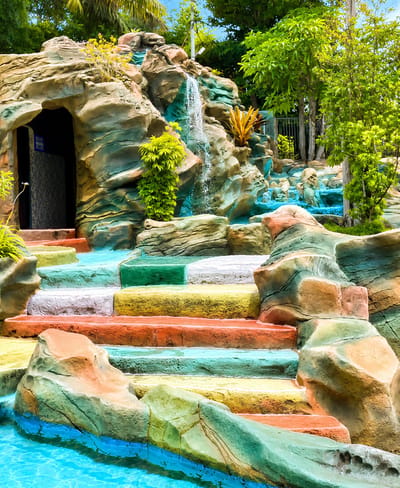 An artificial rock formation with a waterfall and colorful steps leads into a bright blue pool. The rock formation has an opening on the left, resembling a cave entrance, and is adorned with green and brown hues. A small waterfall cascades down the rocks. Brightly colored steps, painted in alternating bands of yellow, orange, white, green, and blue, ascend the rock formation. Lush green trees and foliage surround the scene, with a blue sky visible above. The environment appears to be an outdoor water attraction, possibly a resort or park, located in Eampat Kwalamencirim, Indonesia. There are no people visible in the image.