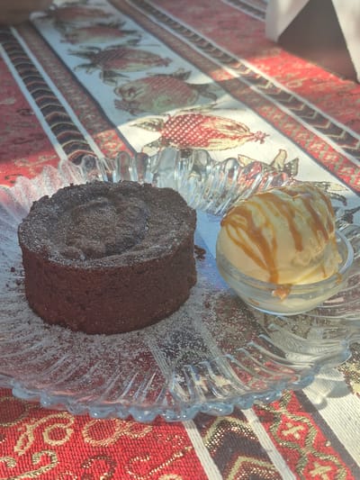 A circular, dark chocolate lava cake is presented on a clear glass plate with a fluted edge. The cake is dusted with powdered sugar, some of which has settled on the plate. To the right of the cake, a small glass bowl holds a scoop of vanilla ice cream drizzled with caramel sauce. The plate and bowl rest on a table covered with a red and white patterned tablecloth. The background displays a runner with a repeating pattern of stylized fish or sea creatures. The scene is lit by natural light, casting shadows across the tablecloth. The image appears to be taken at a restaurant or cafe in Kaluđerac, Montenegro.
