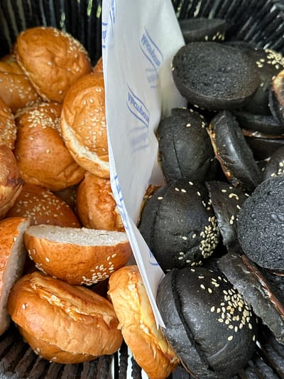 A close-up, overhead shot shows a large number of burger buns. Some are a traditional golden brown color and topped with sesame seeds, while others are a deep black, also sprinkled with sesame seeds. The buns are arranged in a black basket, with a white paper liner separating the two types. The paper has blue text printed on it, likely a brand name. The buns are mostly whole, but some are cut in half, revealing their white interior. The lighting is bright, highlighting the textures of the buns. The scene is devoid of any people or animals. The image appears to be taken indoors, likely in a bakery or food service establishment. The location is Novaleksandrovo, Russia.