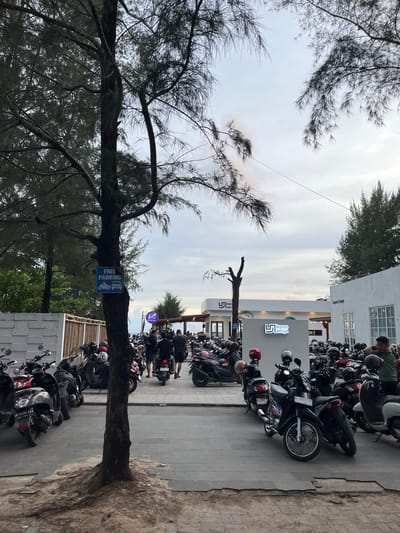 The scene depicts a parking area for motorbikes in what appears to be Monikeun, Indonesia. A large tree stands near the edge of a sandy area, with a blue sign affixed to its trunk indicating "FREE PARKING." The tree's branches reach towards the sky, partially obscuring the view. Rows of parked motorbikes fill the space adjacent to the tree and along the side of a building. The building features a sign with the text "lepas senja" and "Lepas Senja," possibly indicating the name of a business or establishment. People are visible near the motorbikes, suggesting activity in the area. The sky is overcast, with a muted, cloudy tone. The composition is slightly tilted, providing a unique perspective of the location. License plate details such as "BL 4496 AAA" and "EL 4401 PZ" are visible on some of the motorbikes.