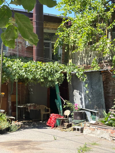 This is a photograph of a sunlit courtyard in Taganrog, Russia. The scene is cluttered with various objects and vegetation, suggesting a lived-in or storage area. In the foreground, a paved surface with some cracks and weeds is visible.
Dominating the mid-ground is a brick building with a metal pipe running vertically along its exterior. A vine-covered structure, likely a pergola or trellis, extends over part of the courtyard, providing shade. Underneath this structure, a table with a floral tablecloth is set with a plastic chair. To the right of this table, another small table and chair are present. A red plastic bag is draped over a bench to the right.
Further to the right, a large, grey, corrugated metal structure is leaning against the building. In front of this, a collection of stacked tires, a large pot containing pink flowers, and various construction materials like cinder blocks are piled up. There are also some green plants growing in the background.
The overall impression is one of a somewhat disheveled but functional outdoor space, likely attached to a residential property. The bright sunlight and clear blue sky suggest a warm day.