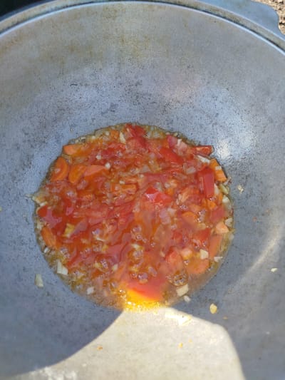 A close-up, overhead shot shows chopped tomatoes and onions sizzling in a large, weathered metal pot. The pot, which appears to be a cauldron, rests outdoors. Sunlight casts a shadow on the lower left portion of the pot, obscuring some of its surface. The ingredients are in the center of the pot, surrounded by oil and juices. Small bubbles are visible around the vegetables, indicating they are actively cooking. The metal of the pot is textured with visible scratches and dark spots, suggesting frequent use and age. No people are visible in the image.