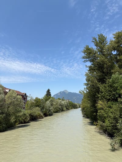 The image depicts a scenic view of a wide, muddy river flowing through a verdant landscape under a clear blue sky dotted with wispy white clouds. Lush green trees line the banks of the river, casting shadows on the water's surface. In the distance, a majestic mountain range rises, its slopes covered in a verdant hue, partially obscured by the trees.
On the left side of the image, a portion of a red-tiled roof building is visible, peeking out from behind the foliage. The sky is a vibrant blue, with a faint contrail from an airplane visible in the upper left quadrant. The overall impression is one of natural beauty and tranquility, suggesting a peaceful, daytime setting. The sunlight filtering through the trees indicates it is likely daytime, possibly late morning or early afternoon. There is no visible text in the image.
The image's orientation is rotated 90 degrees counter-clockwise. The presence of a river, abundant vegetation, and mountains suggests a location with natural waterways and hilly terrain. Given the location context of Cluses, France, this scene is likely a view of the Arve River or a similar waterway in the area, surrounded by the foothills of the French Alps. The weather appears to be clear and pleasant, ideal for outdoor activities or simply enjoying the natural surroundings.