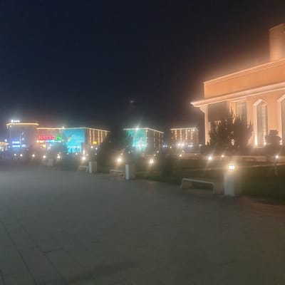 The image shows a nighttime view of several buildings in Turkistan, Kazakhstan. The buildings are illuminated and appear to be commercial establishments. Signage such as "HALVA" and "KFC" are visible. The first building on the very top may be residential. Trees and lamp posts line the street, with a sidewalk visible on the right side of the image. The sky is dark, indicating it is night.