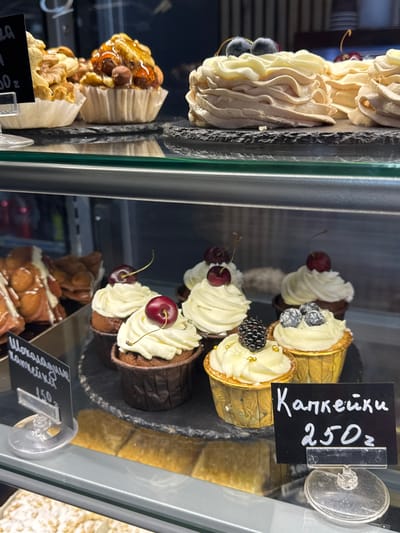 This image displays a close-up view of a refrigerated display case filled with various pastries in a bakery. The subjects are the pastries themselves, which include cream-filled cones topped with cherries and berries, and what appear to be meringues or other light, airy confections. Some pastries are decorated with fresh fruit like cherries and blueberries, while others have golden accents. In the background, a few other baked goods are visible.

The setting is a retail environment, likely a bakery or dessert shop. The display case has glass doors, reflecting the light and surroundings. The lighting appears to be artificial, suggesting an indoor setting, possibly a shop during operating hours.

There is visible text on small black price tags. One tag reads "Капкейки" (Kakeyki - Cupcakes) and "250z". Another tag, partially visible, seems to say "Шоколадный" (Shokoladny - Chocolate) and possibly refers to cupcakes as well, with a price of "150z".

No people or specific activities are depicted, only the static display of the baked goods. The overall impression is one of a well-stocked and appealing confectionery display. The location context provided indicates Alakhadzi, Abkhazia, which suggests the origin or setting of this particular bakery.