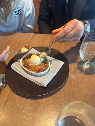 A dessert, likely apple crisp with ice cream, sits on a dark brown plate atop a folded gray napkin. Two individuals are present, each holding a spoon poised to eat. A man in a dark suit with a wristwatch is visible along with a child wearing a gray Santa Catalina sweatshirt. Two water glasses are also visible on the wooden table.