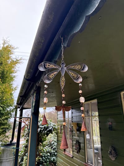 A weathered metal dragonfly wind chime hangs suspended from the dark green eaves of a building in Stiperstones, United Kingdom. The central component is a large, grey-silver metallic dragonfly featuring intricate green-gold patterns within its wings and body. From this, three strands descend, each comprising clear and amber-colored beads, a smaller rustic metal dragonfly, and terminating in a rusty, conical metal bell.
The wind chime is attached via a hook to the underside of a dark green roof overhang, which displays a scalloped edge and a dark, weathered gutter above. The building itself is a dark green wooden structure with vertical siding and multiple windows, reflecting an overcast sky and surrounding vegetation. Several small, rustic metal decorations, including a leaf or bird motif and a pinecone shape, are affixed to the exterior wall of the building. To the left, a dark vertical support column with an attached decorative metal bracket is visible. The background consists of dense green foliage and trees under an overcast sky, with faint utility lines discernible in the upper left. No individuals are present in the scene.