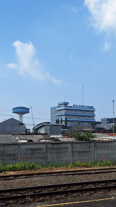 The image shows a cityscape with a large, multi-story building labeled "RS DR. DJOKO PRAMONO" prominently displayed. This building appears to be a hospital or a similar institution, featuring white walls with blue accents on its upper floors and balconies. To the left of this building, a tall water tower with the word "KARAWANG" written on it is visible. The surrounding environment consists of various structures with grey tiled roofs, a concrete wall, and lush green vegetation. In the foreground, railroad tracks are laid across a gravel bed. The sky is a clear blue with scattered white clouds. The scene is captured on a sunny day, with the lighting suggesting it is daytime. There are no people visible in the frame. The presence of the water tower with "KARAWANG" indicates the location is likely within or near the city of Karawang, Indonesia.