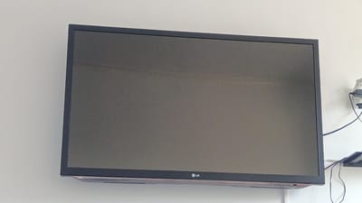 A television screen is mounted on a white wall. The screen is dark, showing no image. The LG logo is visible on the bottom bezel of the television. To the right of the television, a white electrical outlet with a plug and wires is attached to the wall. A small black shelf is mounted below the outlet, with cables draped over it. There are no people in the image. The image depicts a domestic or office setting, common in the city of Maqat, Kazakhstan.