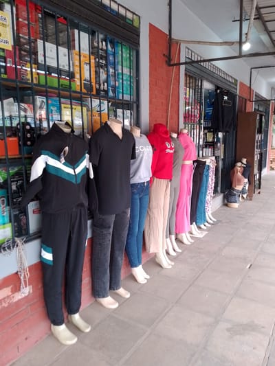 The image shows a row of mannequins displaying clothing outside a shop in Villa Hayes, Paraguay. The shopfront has barred windows displaying various products including electronics. The mannequins are dressed in casual clothing, including tracksuits, t-shirts, hoodies, and jeans. The shop is situated on a paved walkway.