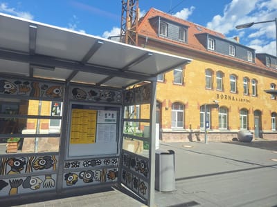 The image depicts a street scene in Borna, Germany, specifically in front of the train station. The foreground features a bus stop shelter with a gray metal frame and a white roof. The shelter's side panels are decorated with colorful, stylized illustrations of faces and abstract shapes. A yellow timetable is displayed within the shelter. To the right of the shelter is a gray trash can.

The background is dominated by a yellow brick building, which is the Borna train station. The building has a red-tiled roof with dormer windows. Above the entrance, the text "BORNA b LEIPZIG" is visible. A streetlamp is positioned to the right of the building. The ground is paved with gray tiles, and there are some shadows cast by the shelter and the building. The sky is blue with scattered white clouds.