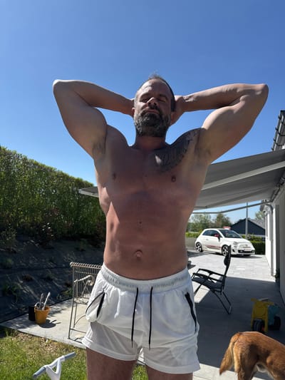 The image captures an outdoor scene featuring a man, a dog, and various outdoor elements under a clear blue sky. The main subject is a man, depicted shirtless from the waist up, wearing white athletic shorts with a black drawstring and a black zippered pocket on the left leg. He has a beard and a large, intricate tattoo on his left shoulder and upper arm. His arms are raised with hands clasped behind his head, and his eyes are closed or squinting, indicating he is either relaxing or stretching in the bright sunlight.
The setting appears to be a paved patio or backyard area, transitioning to a strip of dark ground cover with small plants, and a vibrant green hedge. To the man's right, the hindquarters of a light brown dog are visible. In the background, a white Fiat 500 Abarth car is parked on the paved surface, displaying a Belgian license plate (red characters on white, starting with 'F-'). Other objects include a black metal folding chair, a yellow and blue plastic ride-on toy, and part of a building structure with a retractable awning. A yellow bucket containing cutlery or tools is seen on the ground next to a metal stool.
The activity suggests the man is enjoying the sunny weather, perhaps stretching or simply basking in the sun. The time of day is clearly daytime, characterized by strong sunlight casting defined shadows and a cloudless, bright blue sky. The scene conveys a relaxed outdoor atmosphere. Visible text includes the "Fiat" and "Abarth" branding on the car, and the partial Belgian license plate.