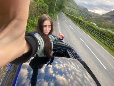 A selfie-style photo depicts a woman with long, dark hair, positioned next to a blue car on a road. She is making a peace sign with her left hand. The car hood reflects the sky with white clouds. The road stretches into the distance, bordered by green fields and trees. Mountains are visible in the background. Based on the visual cues of the landscape, this photo was likely taken near Llangynog, United Kingdom.