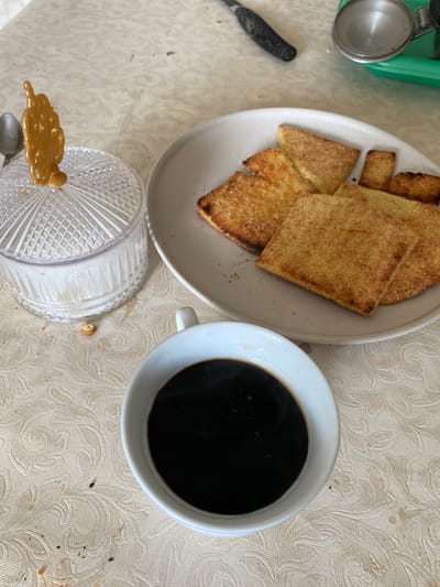 This is a high-angle, full shot of a table setting in Baraki, Algeria, featuring a cup of coffee, a plate of golden-brown, square-shaped crackers or biscuits, and a clear glass jar with a sugar scoop. The table is covered with a light-colored, patterned tablecloth. The coffee is in a white mug, with steam rising, indicating it's hot. The crackers are arranged on a white plate, some overlapping. The sugar jar is topped with a decorative, golden scoop. The background includes a glimpse of a utensil with a black handle and a green tray. The overall scene suggests a breakfast or snack setting.