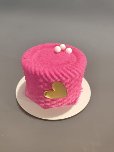 A single, bright pink cake sits on a white round cake board, which is placed on a flat, gray surface. The cake is cylindrical and appears to be frosted with a textured, fluffy pink frosting. There are three small, white, spherical candies or pearls on top of the cake, arranged in a triangular formation. A shiny, golden heart-shaped decoration is affixed to the side of the cake, near the bottom. The background is a uniform, muted gray. The lighting is soft and even, suggesting an indoor setting. No people, text, or weather conditions are visible. The image appears to be a close-up product shot of the cake, possibly for a bakery or online shop.
