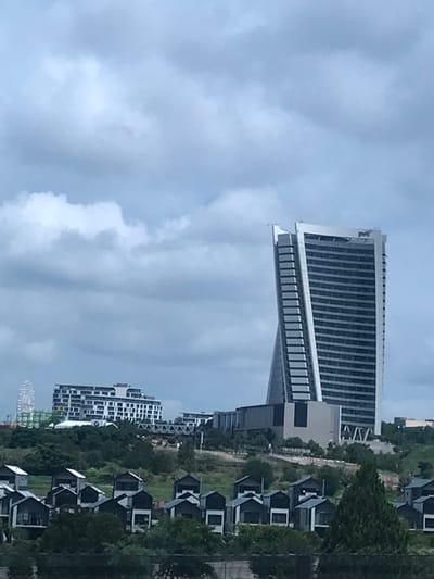 The image presents an urban landscape within Waterfall City, Midrand, South Africa, under an overcast sky. The prominent subject is a tall, modern, multi-story skyscraper, known as the Elliptical Building (or PwC Tower), positioned in the mid-ground to the right. This building features a distinctive, partially twisted or curved architectural design on its facade and displays a "PMF" logo near its peak. To the left, a white, multi-story building with visible balconies, consistent with the Radisson Blu Hotel, Mall of Africa, is situated on a distant rise. Further to the far left, a large white Ferris wheel is partially visible behind the hotel. In the immediate foreground, a cluster of approximately 20 modern residential units are arranged, characterized by dark roofs and light-colored walls, interspersed with green foliage. The overall terrain is undulating and covered with various trees and shrubs. No individuals or specific actions are discernible within the scene.