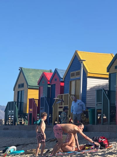 The image shows a row of colorful beach huts against a clear blue sky in Muizenberg, South Africa. The huts are painted in various vibrant colors, including yellow, blue, red, green, and white. Several people are standing near the huts on the beach. A man with a long hairstyle is reaching towards a pile of belongings on the sand. Nearby, a woman in a bikini stands next to him. A young boy with a towel around his neck is standing further down the row of huts. Another man in a light blue shirt is sitting on the steps of one of the huts.