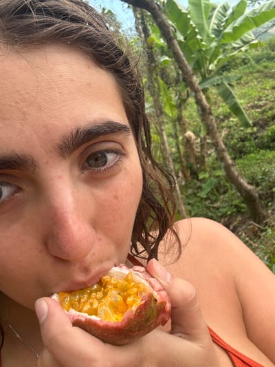 A woman with light skin and brown, curly hair eats a passion fruit. Her face is close to the camera, highlighting her eye color. She wears a rust-colored top. The background features lush greenery, including banana trees, suggesting a tropical location. The image is taken from a slightly low angle.