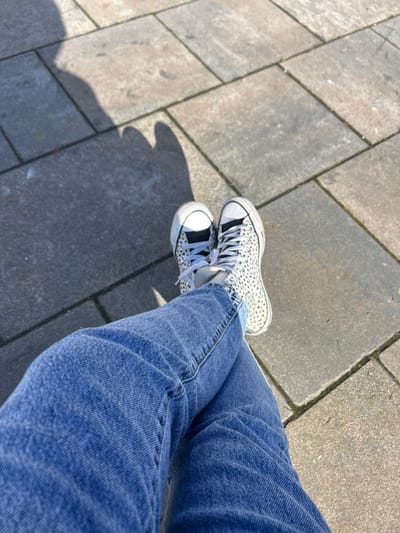The image depicts a first-person, downward-angle view of a person's lower body, situated outdoors on a paved surface. The location is specified as Louviers, France, though no specific landmarks are visible in the frame to confirm this. The individual's legs are visible, clad in blue denim jeans, crossed at the ankles. They are wearing a pair of high-top sneakers featuring a white base with black or dark gray speckles, resembling a dalmatian pattern, and white laces. The soles of the shoes appear white. The ground beneath is composed of rectangular gray paving stones, exhibiting variations in shade and texture. Strong direct sunlight is present, casting distinct, elongated shadows onto the pavement in the upper left portion of the frame, originating from the unseen upper body of the individual taking the photograph. No other people, objects, or interactions are visible.