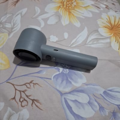The image shows a grey handheld device with a cylindrical handle and a wider, circular head. The head has a black circular lens or opening. On the handle, there are two buttons and a USB-C port. The device is placed on a patterned fabric with floral designs in yellow, white, and purple. The fabric appears to be a tablecloth or bedspread. The background is blurred, but the floral pattern continues. There are no people visible in the image, and no actions or interactions are depicted. The scene is a close-up shot of the device on the fabric. The location of Reudeuep, Indonesia is not visible or suggested by the image content.