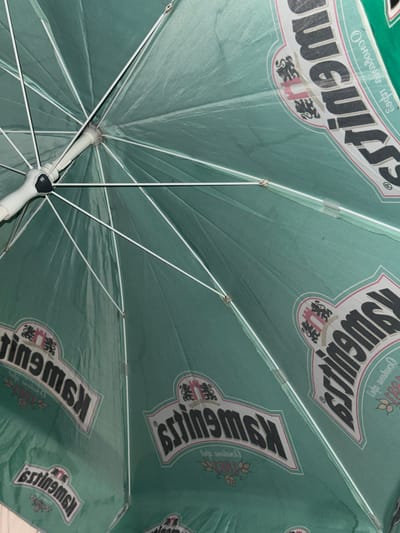 This media file shows the underside of an open green umbrella. The umbrella fabric is patterned with repeated logos. Each logo features a dark, stylized word that appears to be "Estingmak" or "KSWGU!!" in a white and pink border, accompanied by smaller text and what look like crests or emblems with lions. The umbrella's white metal ribs and central mechanism are also visible.

The scene is likely an outdoor setting, possibly in Branipole, Bulgaria, given the location context. The angle suggests the umbrella is held overhead, providing shade. There are no people or animals visible. The lighting is somewhat diffused, but the overall impression is of a bright day, though the specific time of day or weather conditions are not explicitly discernible. The text is primarily in a stylized font for the main logo, with some smaller, potentially Cyrillic script above and below the main words. The repetition of the logo indicates a commercial branding, likely for a beverage or establishment.