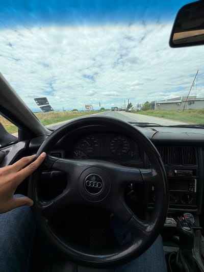 This is a first-person point-of-view image taken from inside a car, likely a vintage Audi, as suggested by the steering wheel emblem. The car is on a paved road, with a slightly cracked dashboard visible. The driver's left hand, wearing a watch and blue jeans, is lightly resting on the steering wheel.
The car is traveling through a rural or semi-rural area, with fields of dry grass or crops lining the road. In the distance, there are some industrial buildings with grey facades and air conditioning units. A billboard is visible on the right side of the road. The sky is a bright blue with scattered white clouds.
The dashboard instruments are visible, showing a speed of approximately 40-50 km/h and a tachometer reading of around 2000 rpm. A radio and gear shifter are also visible in the center console. A pack of cigarettes is on top of the radio. A small orange USB charger is plugged into the cigarette lighter socket. The scene suggests a casual drive through the Bulgarian countryside, possibly near Rakovski, based on the information provided.