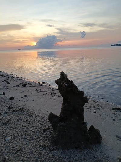 The image depicts a beach scene in Baan Wok Tum, Thailand, during a sunset. The foreground consists of a sandy beach with scattered rocks and debris. A large, dark rock formation stands prominently on the shoreline, partially obscuring the view. Beyond the beach, the water reflects the colors of the sunset. The sky displays a gradient of colors, ranging from light blue to orange and pink, with clouds partially obscuring the sun. The overall scene suggests a tranquil and natural environment.