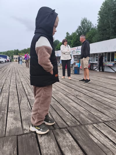 The image captures an outdoor scene on a weathered wooden boardwalk in Nizhnie Yaki, Russia, under an overcast sky. In the immediate foreground, a young blonde-haired child, positioned slightly left of center, faces right in profile, with hands in pockets. The child wears a black hooded jacket featuring small ear-like protrusions on the hood, a cream and light brown fleece mid-section, and matching light brown fleece pants. Dark grey athletic shoes with prominent white soles and yellow accents are visible on the child's feet.
In the midground, a woman with short dark hair, dressed in a light-colored jacket and dark trousers, stands with her arms crossed, appearing to engage in conversation with a man beside her. The man, with short dark hair, wears a dark hooded sweatshirt and light beige shorts, standing with his hands in his pockets. Further back, a series of low white commercial structures extends horizontally across the frame. These buildings feature multiple stalls and display signage in Cyrillic, with "МОРОЖЕННОЕ" (Ice Cream) clearly visible on one sign and "КОФЕ" (Coffee) on another to the right. Several other individuals are observed walking or standing near these structures and further down the boardwalk. Green deciduous trees are visible behind the buildings, forming a natural backdrop. To the far left in the distance, a parking area with several vehicles is present. The wooden boardwalk planks show signs of age and wear.