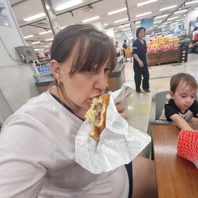 In a well-lit supermarket in Wonju-si, South Korea, a woman is captured in a close-up, mid-bite of a hamburger. She is wearing a white t-shirt and has dark hair with bangs. To her right, a young child sits in a high chair, facing away from the camera, looking down at something on the table. The child wears a black and white shirt. The background reveals aisles of products, including fruits displayed on wooden crates. Other shoppers can be seen in the distance, including a woman in a dark blue top and dark pants, and a person pushing a yellow shopping cart. A sign with Korean text is visible on a shelf. The overall scene suggests a casual outing to a grocery store.