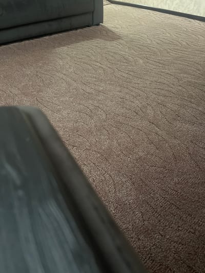 This image shows a close-up view of a textured brown carpet. A portion of a dark gray sofa is visible in the lower left corner of the frame. The lighting suggests an indoor setting, possibly a living room or office space. The image does not provide any specific information to determine its location in Nalchik, Russia.