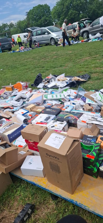 The image captures an outdoor market scene in a grassy field in Gorno Sahrane, Bulgaria, under a partly cloudy sky.
In the foreground, a weathered wooden platform, featuring yellow and blue paint, holds a substantial, disorganized pile of packaged consumer goods. These items are primarily in cardboard boxes and plastic packaging, suggesting new or open-box merchandise. Visible product names include "GyroVu USB Crystal Touch Lamp," "DISwoe," "Wireless Controller For PS4," "3D ILLUSION LAMP," "LED BOXKU," "MP3 Music Player," "USB Cabinet Light," and "Horizontal Employee Badges." Several items display price tags in Bulgarian Lev (лв), such as 20лв, 22лв, 25лв, and 95лв. Black plastic bags and other packaging are intermingled with the products.
The mid-ground features several parked vehicles on the grass, indicating vendor stalls. From left to right, these include a dark grey hatchback with license plate 3671W82, a white commercial van, a silver minivan (likely a VW Sharan) with its rear hatch open, a beige/tan minivan, and a dark grey SUV with its rear hatch also open.
Several individuals are present, engaged in market activities. Near the white van, a person wearing an orange cap and a bright green long-sleeved shirt stands beside a bicycle rack. Another person bends over near the silver minivan's open trunk. A man with grey hair, wearing a light-colored short-sleeved shirt and dark trousers, walks towards the right. Further right, a person is seated on the grass near a spread of clothes and other items.
The background consists of numerous green deciduous trees under a blue sky with scattered white clouds. A dark utility pole or similar structure is visible in the distance among the trees. The overall environment is an open, grassy area characteristic of an informal outdoor market or flea market.