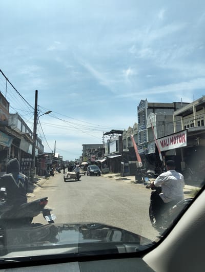 The image captures a street scene in Gampongteungoh, Indonesia, on a bright, sunny day with scattered clouds. The perspective is from inside a vehicle, looking forward. The street is lined with buildings on both sides, appearing to be commercial establishments with awnings and signage. Visible signs include "PLASTIK" and "WALI MOTOR". Various vehicles are present, including motorcycles, a car, and a three-wheeled cart being pulled by a person. People are seen riding motorcycles and walking. The road surface is asphalt. Overhead, a complex network of power lines and a utility pole are prominent. The overall impression is that of a bustling, working street in a town or city.