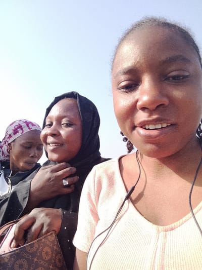 This image depicts three females outdoors under a bright, clear sky in Ibi, Nigeria. In the foreground, a young woman with dark skin and braided hair wears a light peach ribbed V-neck top and wired earbuds around her neck. She appears to be taking a selfie, her mouth slightly open, showing teeth. Behind her, centrally positioned, is a middle-aged woman with dark skin, wearing a black head covering and a dark garment. She is smiling, with her left hand holding her neckline, revealing a ring on her ring finger. Her right hand grips a brown patterned handbag with "CKCA" visible on its surface. To the far left, partially obscured, an older woman with dark skin wears a white garment and a pink and purple patterned headscarf. Her eyes are closed or cast downwards. The lighting suggests bright daylight conditions.
