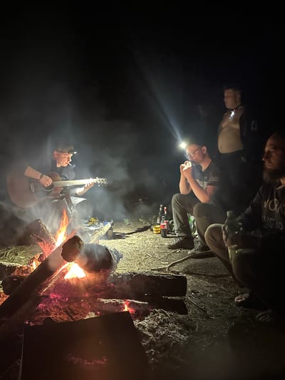 A group of people are gathered around a campfire in a dark, outdoor setting. One person is playing an acoustic guitar while holding a lit cigarette in their mouth. Several individuals sit nearby, one wearing a headlamp. Bottles and other personal items are scattered on the ground. The location is possibly in or near Юзуфоўскі сельскі Савет, Belarus. The scene suggests a casual social gathering at night.