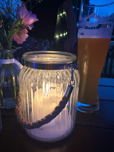 The image depicts a dimly lit outdoor setting, possibly a patio or garden area. In the foreground, a glass hurricane lantern with a thick rope handle contains a lit white candle. The lantern is ribbed and transparent, allowing the candlelight to illuminate its interior. To the left of the lantern, a clear glass vase holds a bouquet of delicate pink and purple flowers, with smaller white blossoms and green stems. A white ribbon is tied around the neck of the vase. To the right of the lantern, a tall glass of amber-colored liquid, likely beer, with a frothy head, stands prominently. The background is dark and out of focus, but suggests the presence of string lights and a person's arm and hand. The overall atmosphere is cozy and relaxed, likely suggesting an evening gathering. The location is identified as Bad Endorf, Germany.