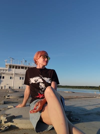 In Kochino, Russia, a woman with short, pink hair and yellow-tinted sunglasses sits on a large, weathered surface. She is wearing a black t-shirt with a graphic design and a denim skirt. Her legs are crossed, and her right hand is resting on her lap. The background shows a large white ship with multiple levels and windows, likely docked or moored. Beyond the ship, a calm body of water stretches to a tree-lined horizon under a clear, bright blue sky, suggesting a sunny day, possibly late afternoon due to the warm lighting and long shadows. The overall scene evokes a sense of relaxed contemplation in an outdoor, waterfront environment.