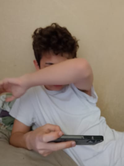 In an indoor setting, a person with curly dark hair is lying down and holding a smartphone. The individual is wearing a white t-shirt, and their arm is covering their face. The background appears to be a plain wall. There is no indication of the city of Motyakovo, Russia, or any other specific geographical location.