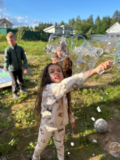 In Oseyevo, Russia, on a sunny day, two children are playing in a grassy backyard. A young boy, with blond hair, stands in the background wearing a dark green jacket and black pants, holding a green bubble wand. In the foreground, a young girl with long dark hair is in focus, her face turned towards the camera. She is wearing a light-colored pajama set with a pattern of cats and dogs. She is actively blowing bubbles, with a cluster of large, shimmering bubbles suspended in the air in front of her. Her right hand is holding a white bubble wand with a green loop. The environment is a rural setting with houses and trees visible in the background, under a bright blue sky with scattered clouds. A soccer ball is visible on the ground to the right of the girl.