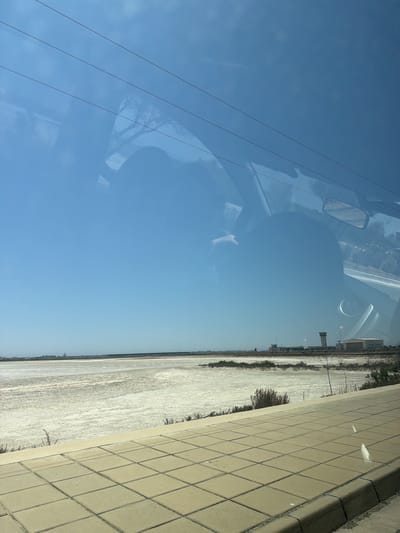 The image, taken from a vehicle in Larnaca, Cyprus, shows a reflection on the windshield. The reflection includes two indistinct human heads, possibly of the occupants of the vehicle, and the silhouette of a tree. In the background, a vast, dry, salt-covered landscape stretches to the horizon under a clear blue sky. A paved walkway or roadside is visible in the foreground, and in the distance, several structures, including what appear to be airport buildings and a control tower, are present. The overall scene depicts a bright, sunny day with a clear view of the surrounding environment, with reflections obscuring parts of the image.