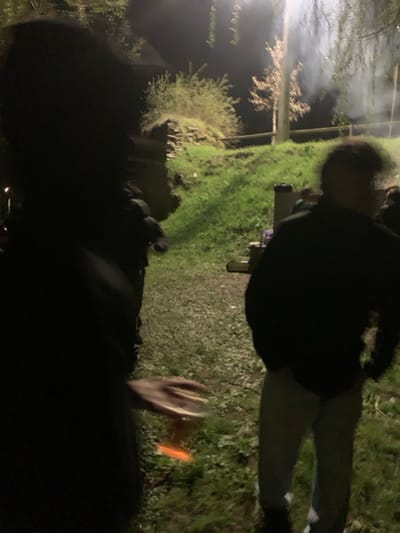 This is an outdoor nighttime scene in Macedo de Cavaleiros, Portugal, illuminated by artificial light. In the immediate foreground on the left, a dark, largely silhouetted and out-of-focus human figure is visible. A right hand, blurred by motion, extends from this figure, holding a clear glass containing an amber liquid. To the right of the frame, a second human figure stands, viewed from behind, wearing a dark top and light-colored trousers; this figure's head is blurred. The ground beneath the figures consists of a gravel path transitioning to an area of vibrant green grass, both illuminated. In the midground, a dark object resembling a cooler or small cabinet rests on the grass. The background features a grassy incline with a low stone structure partially covered by vegetation. Several trees are present, including one with bare branches, and smaller bushes. A plume of white smoke or mist rises from the right background, partially obscuring vegetation. A dark, indistinct building is faintly visible in the far left background.