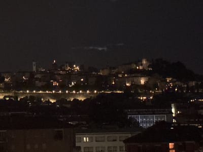 The image shows a nighttime view of the city of Bergamo, Italy. The scene is dominated by darkness, with the lights of the city visible in the distance and in the foreground. In the background, on a hill, are the lights of the Città Alta, the upper town of Bergamo, with some buildings and possibly a tower illuminated. In the middle ground, there appears to be a line of lights along a street or embankment, with trees and darker buildings interspersed. In the foreground, several buildings are visible, some with lit windows. The overall impression is of a cityscape at night, with a focus on the illuminated structures against the dark sky.