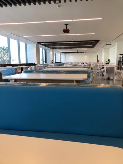 The image depicts a large, modern dining hall or cafeteria located in Hanak, Saudi Arabia. The expansive interior features numerous tables, chairs, and booth-style seating arrangements, all predominantly covered in clear plastic sheeting, indicating either new installation, protection during renovation, or preparation for opening.
The foreground is dominated by a blue booth backrest with a beige tabletop in front of it. Rows of wrapped tables, with white tops and light-colored bases, and chairs, also wrapped in plastic, extend into the midground and background.
The room is brightly illuminated by natural light entering through large, vertical windows on the left wall, which offer a glimpse of outdoor trees and a bright sky. Additional illumination comes from numerous linear LED light fixtures embedded in the white ceiling. The ceiling also features dark brown slatted sections, a white smoke detector, and a centrally suspended black digital clock displaying "1:13" in red.
Towards the far right of the room, a single individual is visible, seated at a table. This person wears a high-visibility yellow or lime green vest over dark clothing and is engaged with a mobile device. Further right, a black beverage dispenser unit is situated on a counter. Multiple red fire extinguishers are mounted on the white walls throughout the space. A projector is mounted on the ceiling in the midground, right side. The flooring is a light-colored, uniform surface.